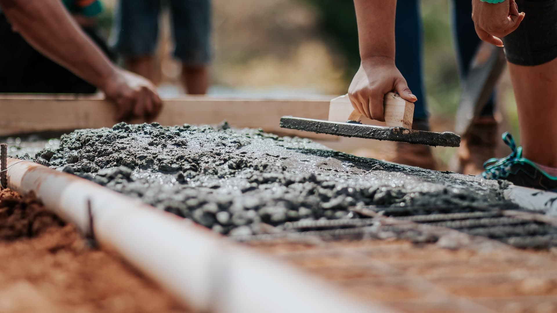 Mains qui lissent du béton frais avec un outil à manche en bois, personnes floues en arrière-plan